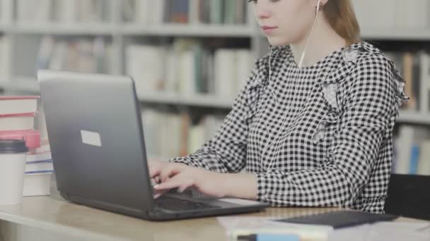 Schöne Studentin mit Kopfhörern sitzt am Schreibtisch in der Bibliothek und arbeitet konzentriert an Schulprojekt. — Stockvideo