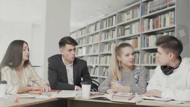 Dos hombres y dos mujeres estudiantes universitarios riendo mientras se preparan para los exámenes mientras están sentados en la mesa de la biblioteca universitaria — Vídeos de Stock
