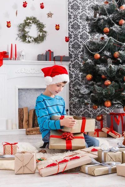 Lindo chico en santa sombrero desenvolver regalos de Navidad —  Fotos de Stock