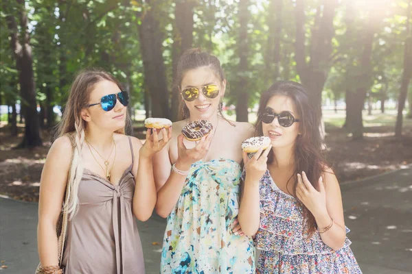 Three beautiful young girls eat donuts in park. — Stock Photo, Image
