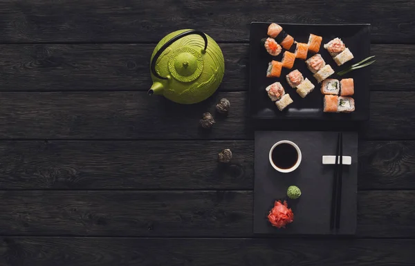 Set of sushi and rolls with teapot on black rustic wood, top view — Stock Photo, Image