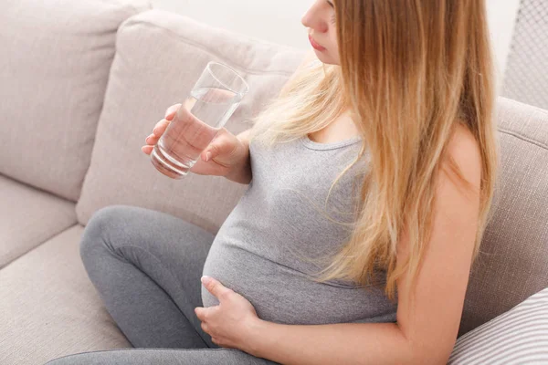 Gravid kvinna med glas vatten sitter på soffa — Stockfoto