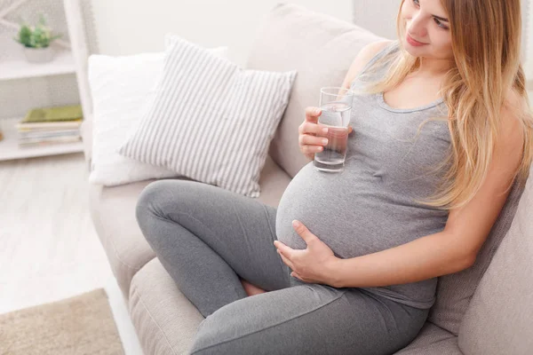 Kobieta w ciąży, popijając szklanką wody, siedząc na kanapie — Zdjęcie stockowe