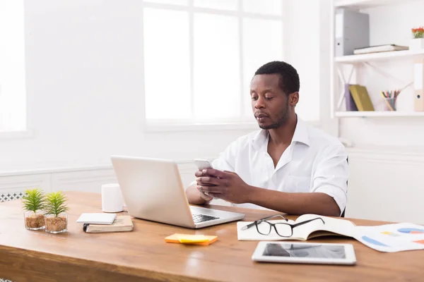 Jonge Afro-Amerikaanse zakenman texting op telefoon terwijl u werkt op laptop — Stockfoto