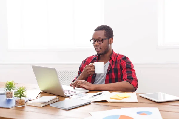 Homme d'affaires noir détendu dans un bureau décontracté, travailler avec un ordinateur portable, boire du café — Photo