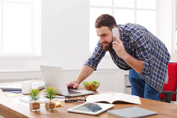 Молодой бизнесмен, стоящий у ноутбука и разговаривающий по телефону в современном белом офисе — стоковое фото