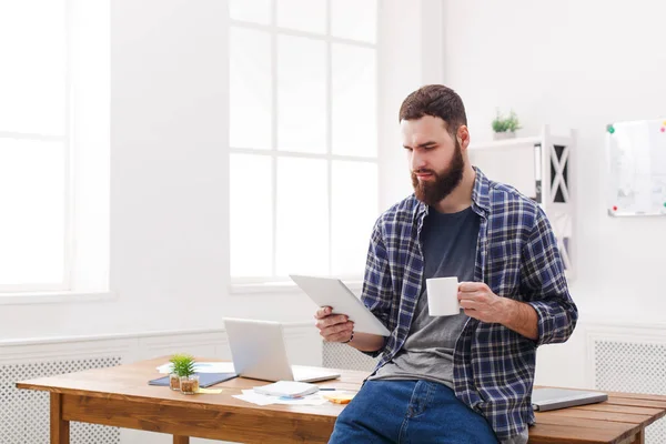 Lässig sitzt ein junger Mann mit einer Tasse Kaffee am Schreibtisch und benutzt ein digitales Tablet im modernen Interieur des Arbeitsplatzes. — Stockfoto