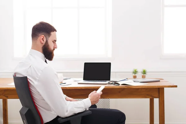 Молодой успешный бородатый бизнесмен в рубашке с ноутбуком и мобильным телефоном в современном белом офисе . — стоковое фото