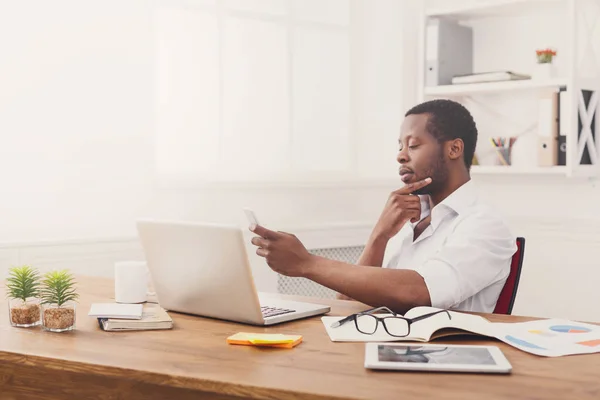 Doordachte Afro-Amerikaanse zakenman texting op telefoon terwijl u werkt op laptop — Stockfoto