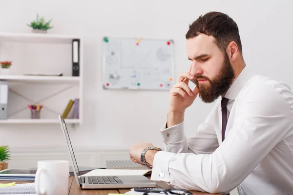 Hombre de negocios pensativo en camisa blanca mirando el reloj hablando en el móvil con el ordenador portátil. Vista lateral — Foto de Stock