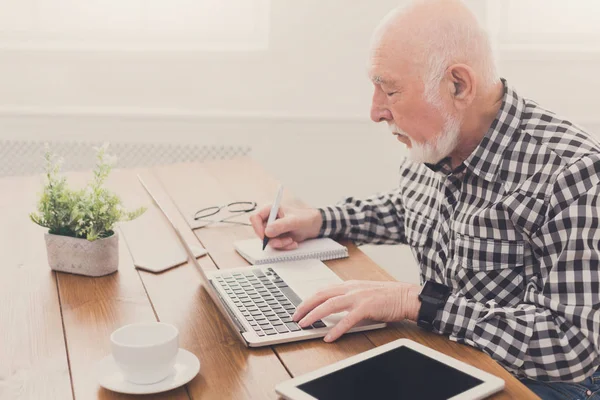 Reifer Mann benutzt Laptop und schreibt in Notizblock — Stockfoto