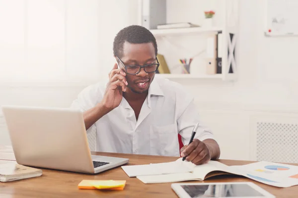 Jonge zwarte zakenman maken van aantekeningen en praten op mobiele in moderne witte kantoor — Stockfoto