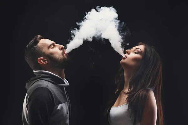 Молодая пара, смочивающая электронную сигарету с дымом на черном крупном плане — стоковое фото