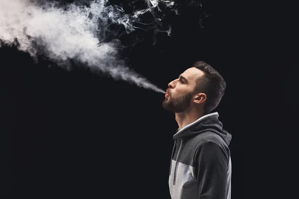 Hombre joven vapeando e-cigarrillo con humo en negro — Foto de Stock