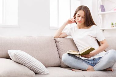Genç esmer kadın evde kanepede kitap okuma.