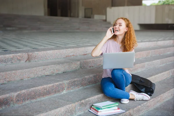 Cep telefonu, belgili tanımlık merdiven açık havada oturup konuşurken laptop ile çalışan genç redhead öğrenci kız