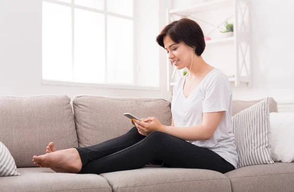 Mujer joven pensativa con teléfono y auriculares en sofá beige —  Fotos de Stock