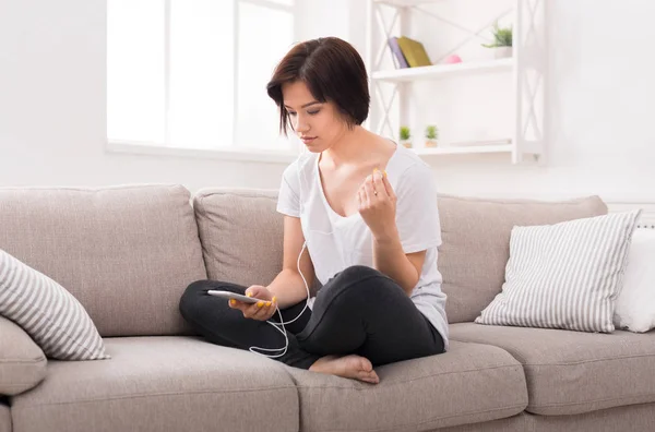 Nachdenkliche junge Frau mit Telefon und Kopfhörer auf beigem Sofa — Stockfoto