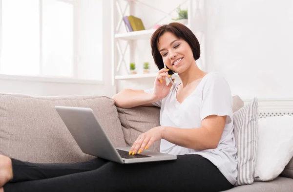 Jovem sorrindo mulher bonita com telefone e laptop no sofá — Fotografia de Stock