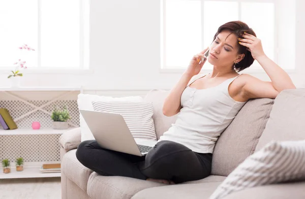 Pensive girl with laptop and mobile sitting on beige couch — Stock Photo, Image