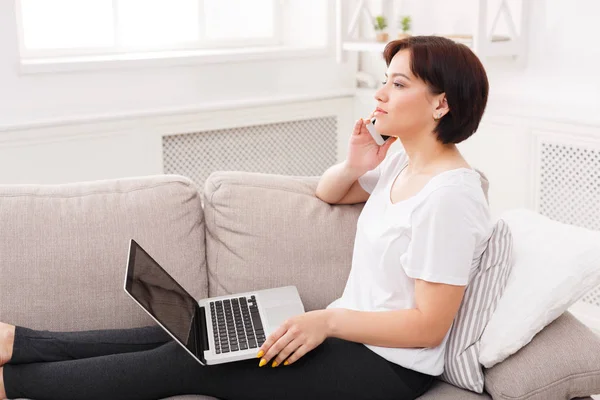 Junge nachdenkliche schöne Frau mit Telefon und Laptop auf dem Sofa — Stockfoto