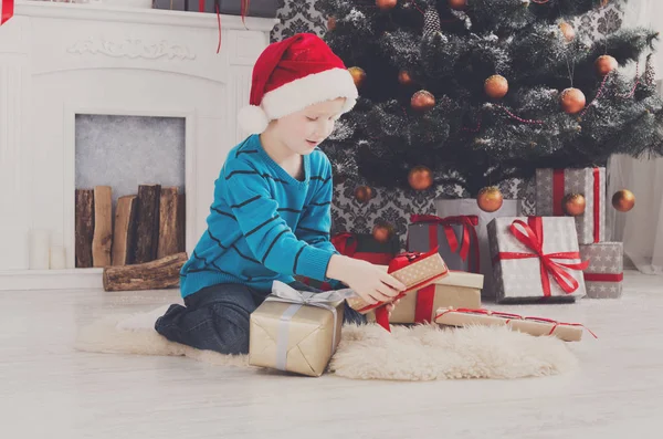 Menino bonito em santa chapéu desembrulhando presentes de Natal — Fotografia de Stock