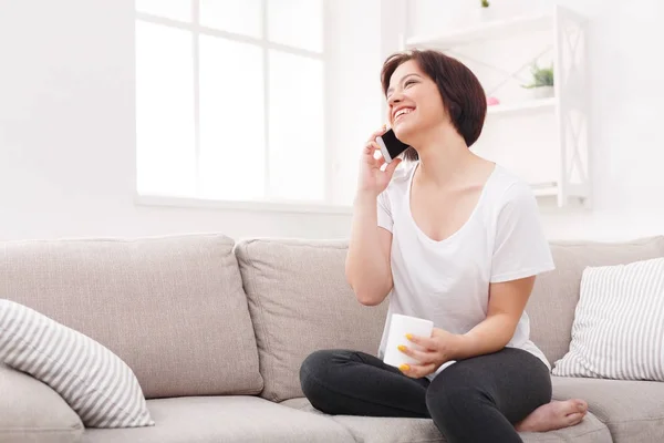 Усміхнена дівчина розмовляє по смартфону і п'є каву вдома — стокове фото