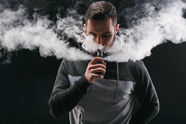 Young man vaping e-cigarette with smoke on black — Stock Photo, Image