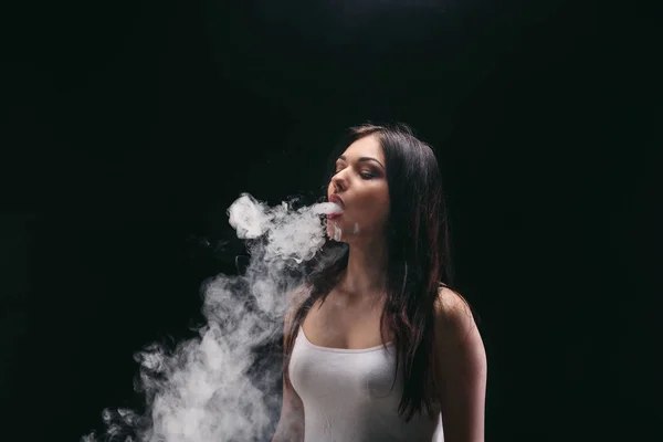 Молодая женщина, испаряющая электронную сигарету дымом на черном — стоковое фото