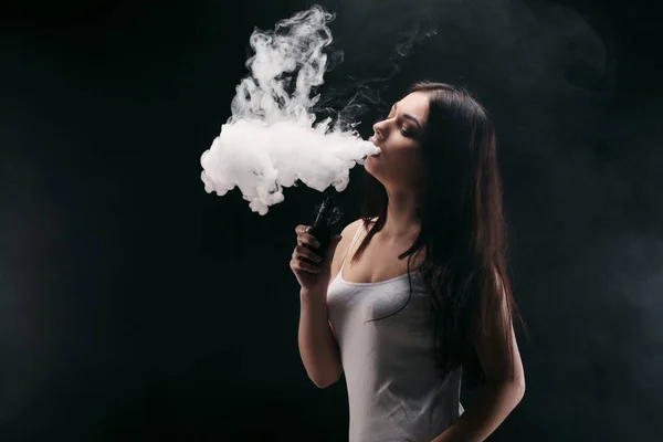 Молодая женщина, испаряющая электронную сигарету дымом на черном — стоковое фото