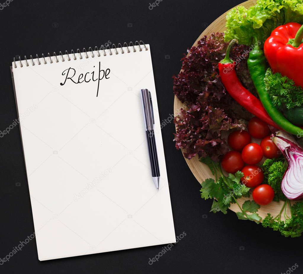 Notepad for ingredients list and big platter with fresh vegetables on black background. Mockup for healthy dishes recipes