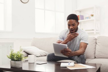 Şaşırmış genç adam evde tablet üzerinde okuma