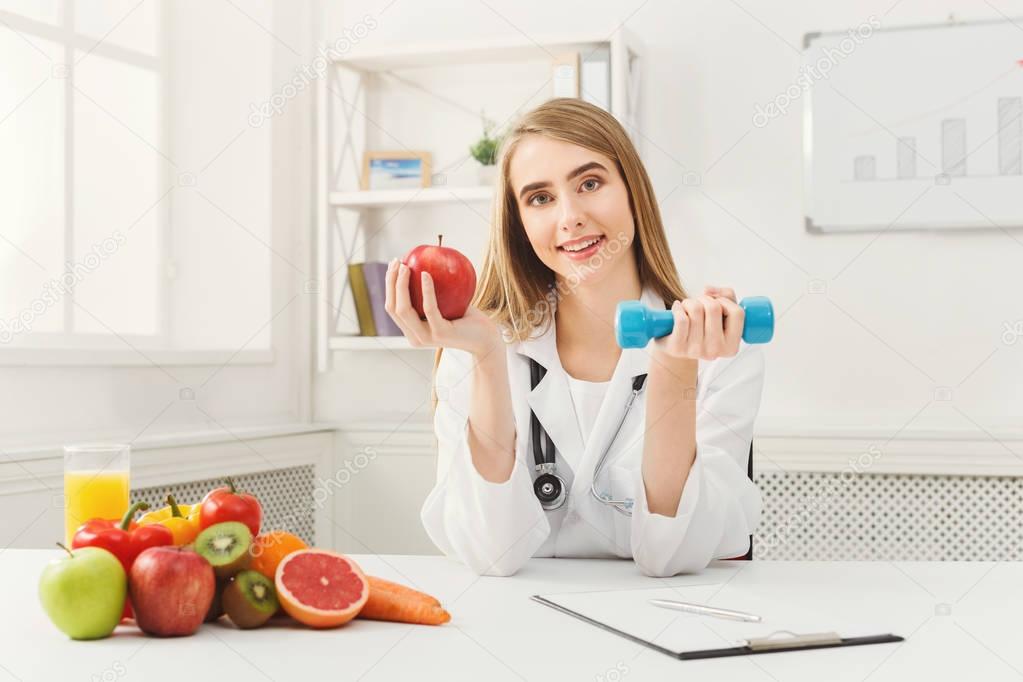 Young dietitian with apple and dumbbell at clinic