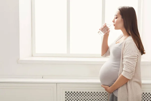 Pensativo mujer embarazada beber agua cerca de la ventana —  Fotos de Stock