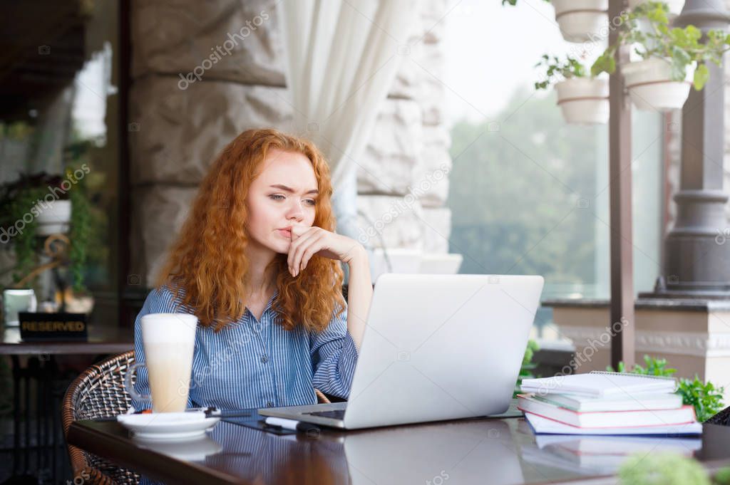 Young pensive redhead girl looking at laptop sitting outdoors