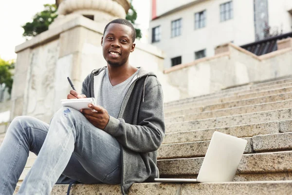 Χαμογελώντας Αφρικανικός-Αμερικανός φοιτητής που σπουδάζει με laptop και μικρόs φορήτοs σε εξωτερικούς χώρους — Φωτογραφία Αρχείου