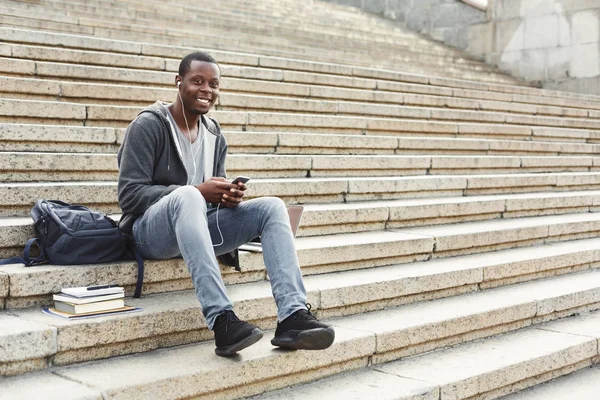 African-american student sitting on stairs and using his smartphone outdoors