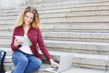 Merdivenlerde oturan, notlar okuma ve dizüstü bilgisayar açık havada kullanarak öğrenci kız