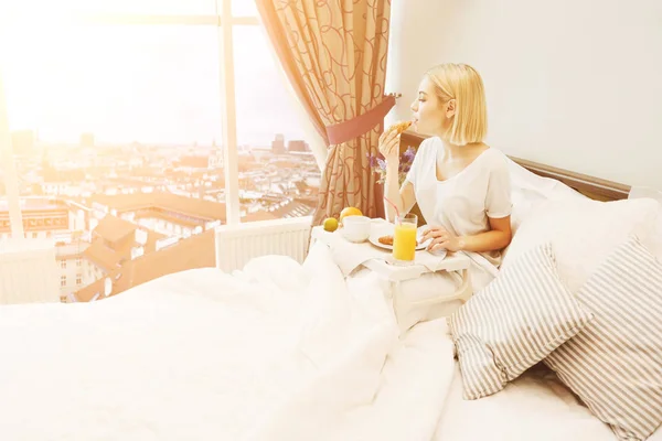 Schöne junge Frau sitzt auf dem Bett und frühstückt — Stockfoto