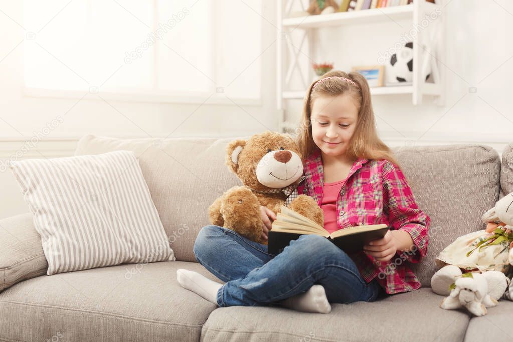 Happy little female child hugging her teddy bear and reading book on sofa at home