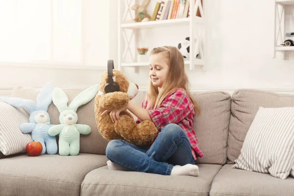 Menina pequena feliz e seu ursinho de pelúcia ouvindo música no sofá em casa — Fotografia de Stock