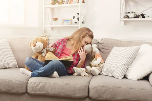 Happy little female child hugging her teddy bear and reading book on sofa at home — Stock Photo, Image