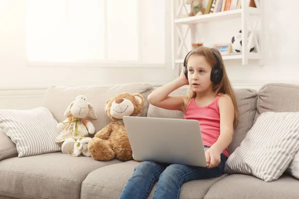 Aranyos kis lány csinál házi feladatot, laptop, fejhallgatót a fotelben ülve — Stock Fotó
