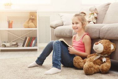 Mutlu küçük kadın çocuk ve onun oyuncak ayı yerdeki evde kitap okuma