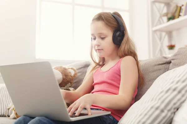 Roztomilá holčička dělá domácí práce na notebooku, sedící na gauči v sluchátka — Stock fotografie