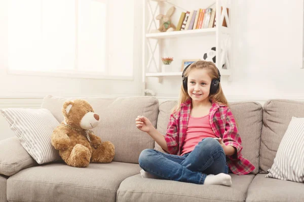 Happy malá holčička a její teddy bear, poslech hudby na gauči doma — Stock fotografie