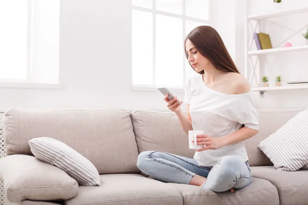 Молода жінка сидить на дивані з мобільним телефоном і кавою — стокове фото