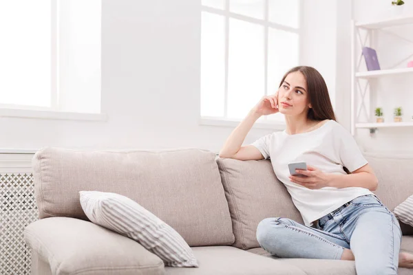 Mujer joven pensativa sentada en un sofá con teléfono celular —  Fotos de Stock