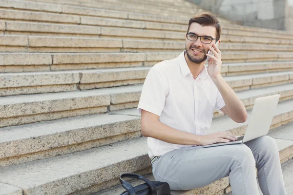 Joven empresario alegre usando el ordenador portátil y haciendo una llamada en sta — Foto de Stock