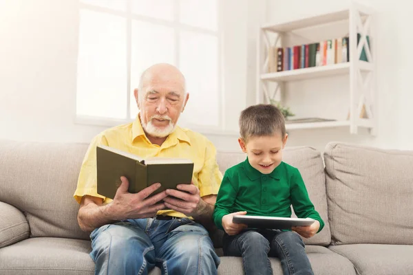 Дідусь читає книгу, онук використовує планшет — стокове фото